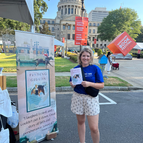 Hickner poses with her book at the Mississippi book festival. Her marketing banner is to the left, the Mississippi capital and the Mississippi Book Festival statue are behind her