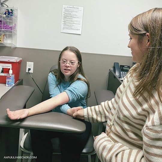 photo of Penny sitting in a chair in a doctor's office. She is waiting to get her blood drawn and Amy Julia is standing by her holding her hand.