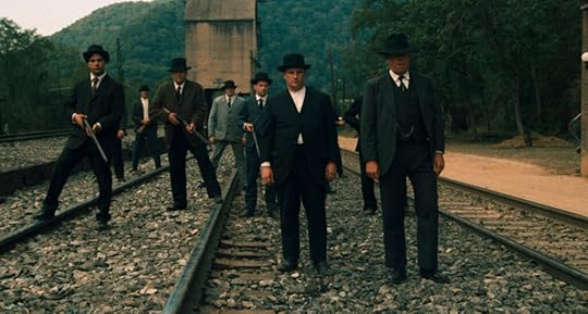 Several men in suits and hats of the early 201th century stand across a set of railroad tracks.
