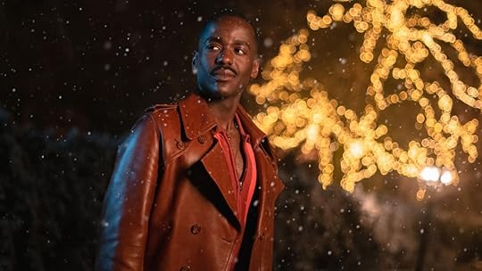The Fifteenth Doctor stands outside in the snow with a tree lit up behind him
