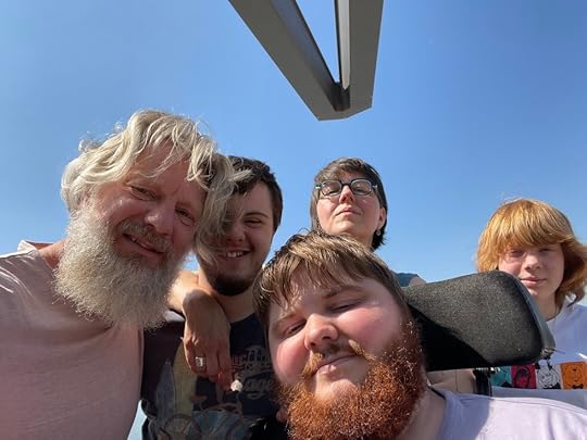 Een familieportret met vijf mensen die samen poseren onder een heldere blauwe lucht. De groep bestaat uit een man met een baard, een jonge man met kort haar en een andere jonge man met een volle baard, een vrouw met een bril en een jongen met rode haren.