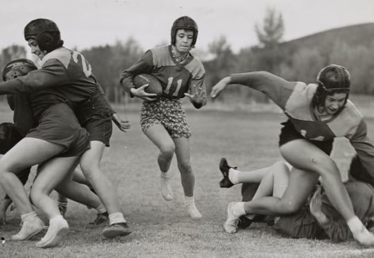 Black and white photo of handful of girls, wearing shorts, numbered jerseys and leather helments, in the middle of a football play, with #11 running the ball toward the camera