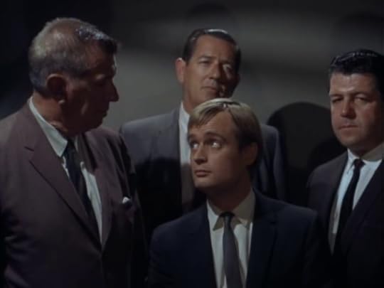 Illya Kuryakin in an elevator with three large men. He looks up at one of them.