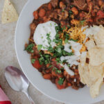 white bowl of chili with toppings and chips, a spoon on the table nearby.