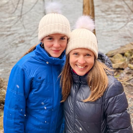 Marilee and Amy Julia smile at the camera. They are wearing winter coats and matching white stocking hats and are standing in front of a river.