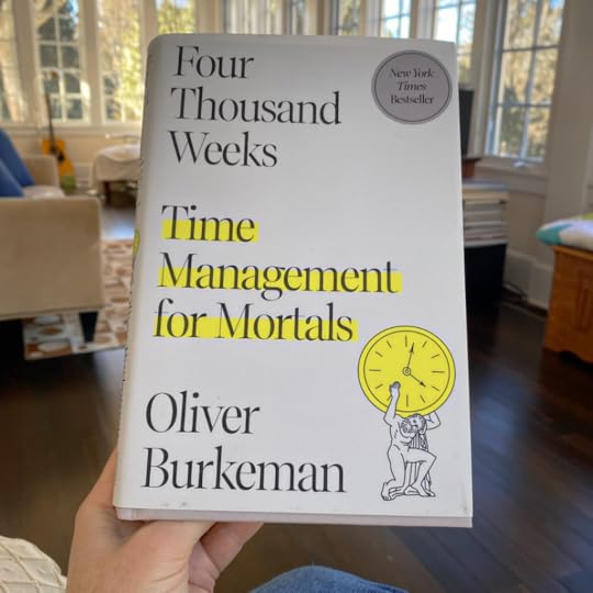 A hand holds up the book Four Thousand Weeks (the book cover is white with the title and author's name, Oliver Burkeman, in black text, with the words 