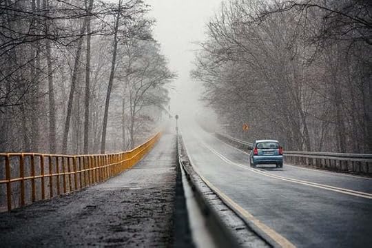 Snowy road with a car traveling away from viewer on the right, a sidewalk on the left, and a high yellow rail between sidewalk and trees.