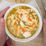 hands gripping a bowl of chicken noodle soup.
