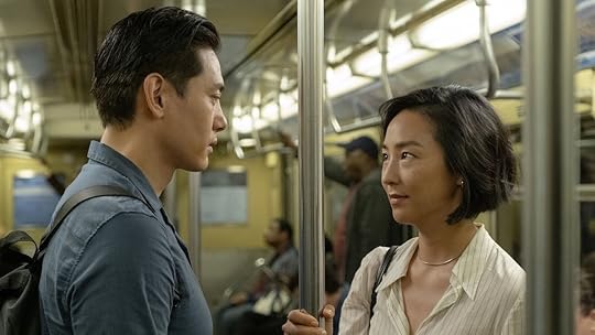 A man in a blue shirt looks at a woman holding the pole on a train in Past Lives
