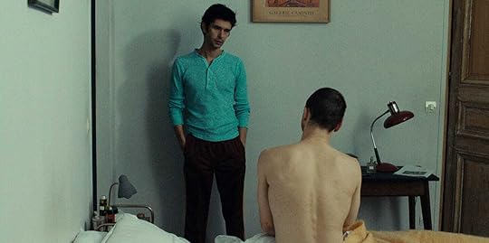 Clothed Ben Whishaw stands next to a nude, seated Franz Rogowski in