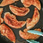 teal tongs picking up skillet chicken tenderloin heavy with seasoning.