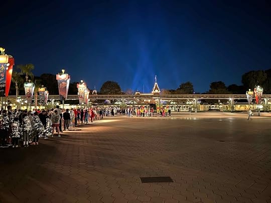 A line of people are waiting to go through the turnstiles at the front of Disneyland. Floodlights are highlighting the castle in the background.