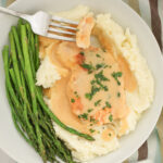 a bite of chicken on a fork over a plate of chicken and gravy with asparagus on the side.