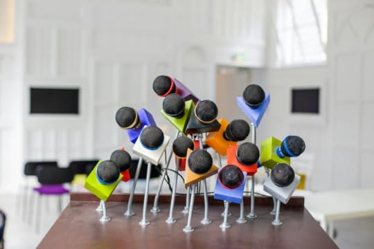 Image: in preparation for a press conference, numerous microphones are attached to a dais, each embellished with a brightly colored triangular or square block to differentiate them.