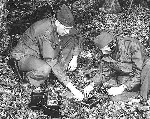 Two men in uniform working on a radio in the woods.