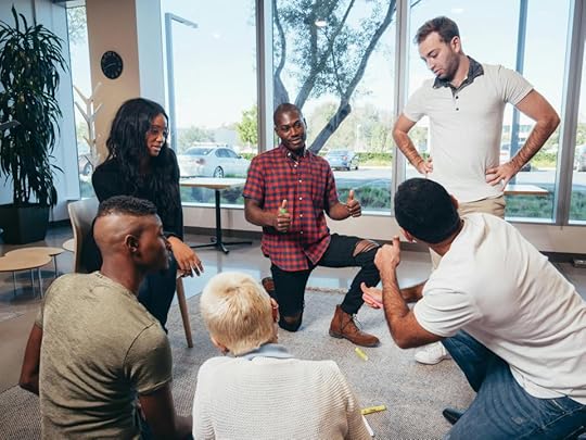 A diverse group of six people engaged in a discussion or brainstorming session in a modern office space, seated in a circle on a carpet. One person is animatedly speaking while others listen attentively.