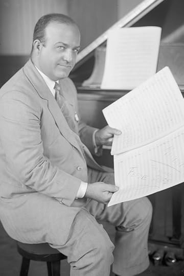 Ferde Grofé, wearing a suit and tie and seated on a piano bench, half-turned to the camera, holds two pieces of sheet music