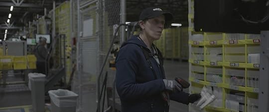 Frances McDormand in an Amazon baseball cap standing in front of pallets in an Amazon warehouse