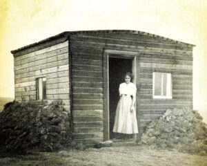 woman in long dress stands outside door of simple frame dwelling, a typical Montana shack occupied by homesteaders