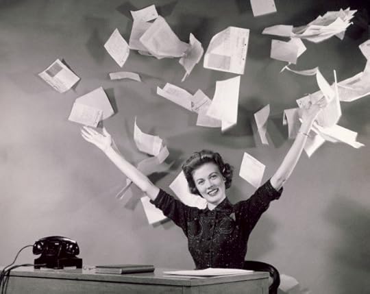 Black and white photo from 1960s of woman seated at desk on which is an old telephone, a book and some papers. She is smiling and holding her hands above her head, having just tossed a stack of papers up into the air.
