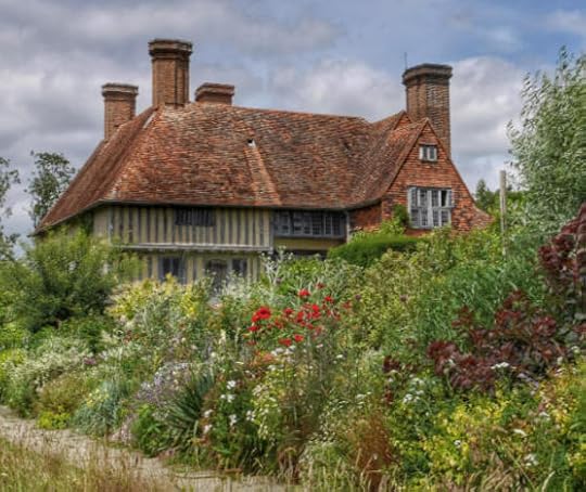 A Tudor style house with a tall gable roof and four prominent chimneys, set back behind a lush English garden of flowers, trees, and shrubs.