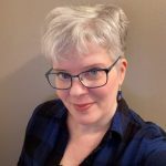 A close-up of me, standing in front of a beige background. I am a white woman with short silver hair cropped closely on the sides. I am wearing dark teal metallic rimmed glasses with rhinestones on the side. I have blue-green eyes, coral lipstick, and I’m wearing silver hook earrings with black and electric blue beads. My shirt is plaid with black and electric blue.