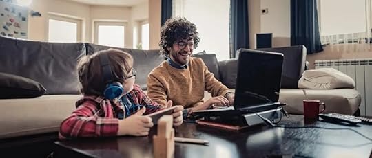 Father working on a laptop next to his son who is wearing headphones and playing on a tablet. This photo highlights the need for self-compassion for parents