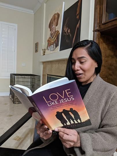 A woman sitting in a cozy living room, reading the book 'Love Like Jesus,' surrounded by decorative art on the walls.