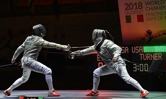 Two sabre fencers on an elevated strip in spotlighting with a large scoring machine behind them. The name on the scoring machine is