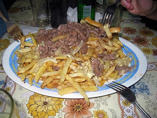 Chorrillana - a plate of fries with cut up meat on top, with three forks reaching out for it, set on a table with bottles in the background.