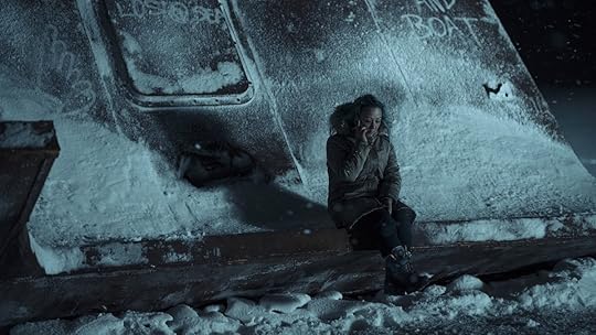 A woman bundled up in a winter coat on her cellphone sitting against a ruined boat covered in ice in true Detective: Night Country