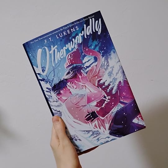Photo of Otherworldly by FT Lukens. Cover art is two teens sort of falling into each other and looking like they're maybe about to kiss, in a snowy setting. The supernatural has a glowing yellow eye and is wearing short-sleeves, unbothered by the cold. The human is bundled up. The people are colored with lots of pink and purple, the background with white and blue.