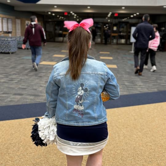Penny, dressed in her blue and white cheerleading uniform and wearing a jean jacket over it and a pink bow in hair, walks down the school hallway away from the camera