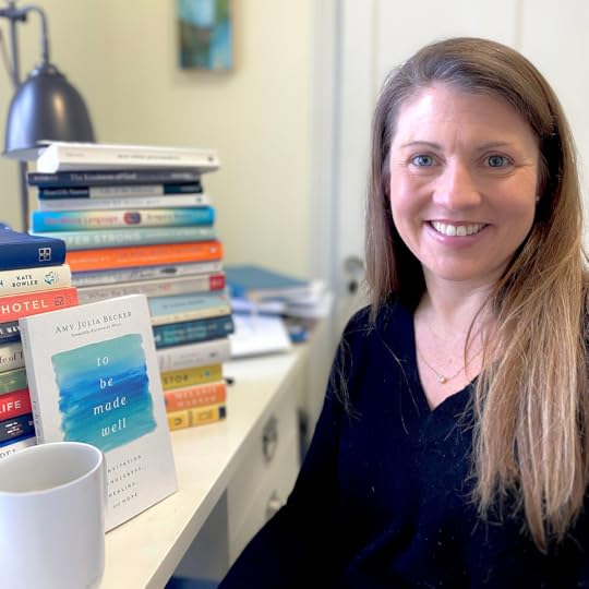 photo from 2022 of Amy Julia, who smiles at the camera and sits at a desk. On the desk, the book To Be Made Well is propped up in front of stacks of books next to a white mug