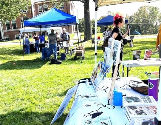 A table outdoors with books and bookmarks on it, with popup canopies int the background over tables of books, and people milling about.