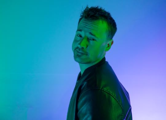 A man is illuminated in blue and green and stands with his shoulder to camera while looking into the camera with a smirk