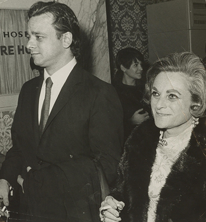 Half-lenth portrait of a man and woman in formal attire, with linked arms, walking into a room. 