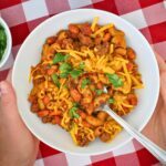 hands holding a white bowl of chili mac with a spoon and toppings.