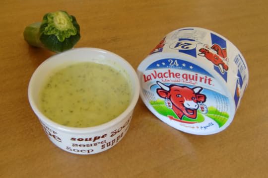 A dark green zucchinni rests on a bowl of soup, filled with light green soup, next to a round box of 24 triangles of Laughing Cow cheese.