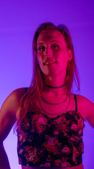 A woman with long brown hair stands akimbo, illuminated in red with a purple backdrop