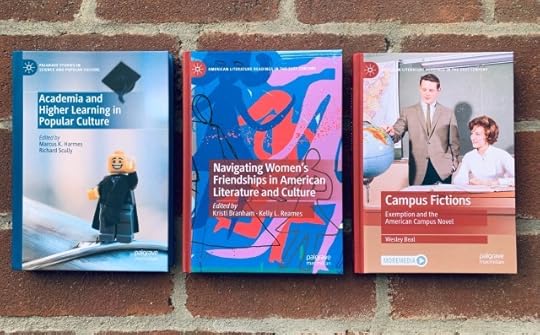 Three hardback books are lined up on a brick walk: Academia and Higher Learning in Popular Culture (2023, with cover art of a Lego graduate throwing his cap into the air), Navigating Women's Friendships in American Literature and Culture (2023, with cover art of abstract women's figures), and Campus Fictions: Exemption and the American Campus Novel (2024, with cover art depicting a vintage classroom with two retro figures looking at a globe).