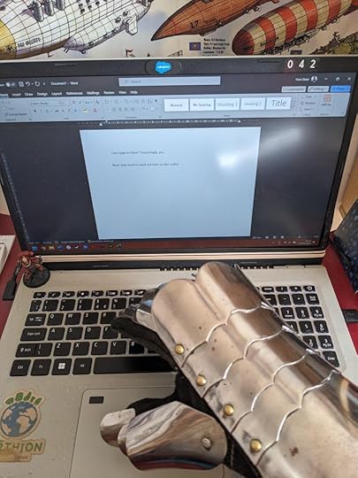 A gauntleted hand typing at a laptop keyboard. The screen reads: 'Can I type in these? Surprisingly, yes. Now I just need to work out how to take a pho-'