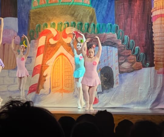 Penny dances on stage with a scene from the Nutcracker behind her. She and two other dances wear tutu dresses and have their arms rounded and raised above their heads