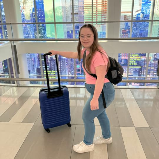 Penny stands in a terminal with colorful buildings out the window. Her hand is guiding a suitcase