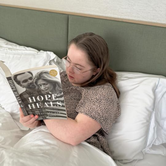Penny is under the covers at a hotel and is reading the book Hope Heals
