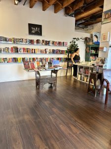 Inside the Maktaba bookshop. 3 shelves on the wall to the left, filled with books facing out. There is a counter to the right with one of the owners standing beside it wearing a black tank top and black pants and white sneakers. A brown hardwood floor at the front.
