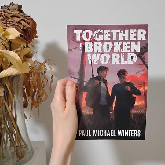 Photo of my hand holding Together in a Broken World by Paul Michael Winters, next to some dried roses. The cover is art of two teen boys carrying packs, electricity posts and wires behind them, all against a pink and orange sunset.