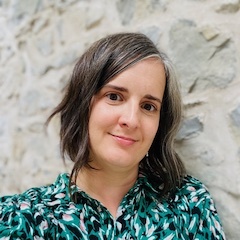 A smiling light-skinned woman with medium length hair and a colourful green top in front of a stucco wall.