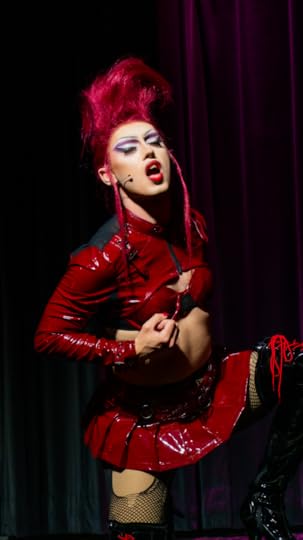 A drag queen sits on a stool with arms outreached