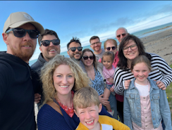 A family photo - nine adults and three children huddled together on a beach. It's windy, their hair is blowing, and everyone has long sleeves. Despite the seeming cold, everyone is grinning and looking happy.